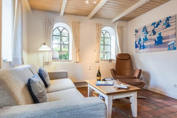 A cozy living room with a sofa, a wooden coffee table, and a brown leather chair. There are two windows with curtains, a lamp, and a painting on the wall.