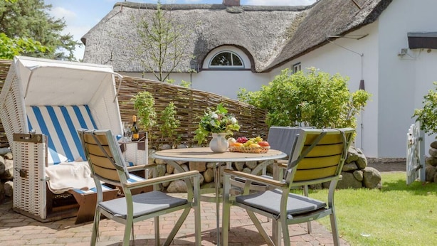 A patio with a round table and four chairs, a wicker chair, and a thatched-roof building in the background. - Ruhig. Edel. Garten. Haus Mühlenhof, Whg. 4 Objekt-id: Mht/04 (Westerland)