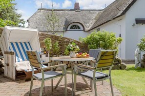 A patio with a round table and four chairs, a wicker chair, and a thatched-roof building in the background.