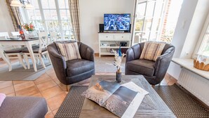 Un salón con dos sillones de cuero, una mesa de centro con revistas, un televisor sobre un mueble bajo y una zona de comedor con una mesa y sillas.
