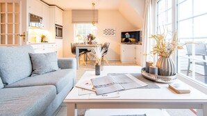 Ein modernes Wohnzimmer mit grauem Sofa, einem Essbereich mit Tisch und Stühlen sowie einer Küche mit weißen Schränken und Geräten.