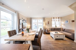 En rummelig opholdsstue med spisebord, sofa og store vinduer der giver udsigt til naturen.