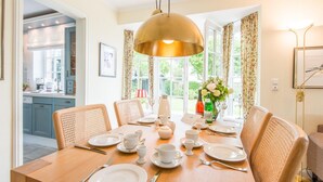 A dining area with a wooden table set for four, surrounded by wicker chairs. A brass pendant light hangs above the table, and a vase with flowers is on the windowsill. - Behaglich. Britisch. Zuhause (Westerland)