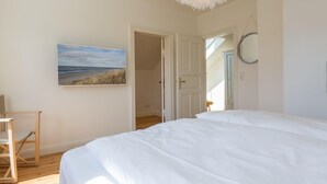 A bedroom with a large bed, a wooden chair, a painting of a beach, and a white door leading to another room. - Bäderstil. Strand. Sauna. Objekt-id: Ebw/48 (Westerland)
