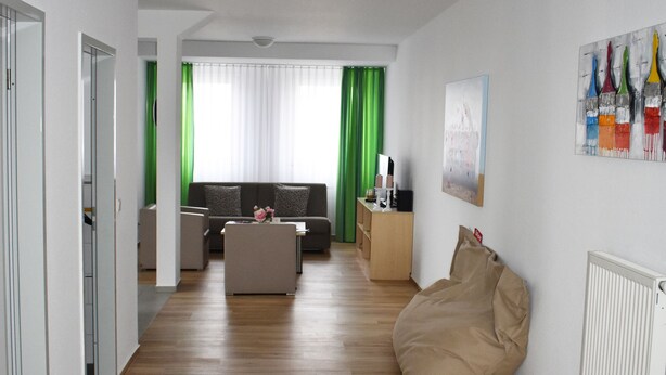 A modern living room with a sofa, coffee table, and a bean bag chair. There is a dining area with a table and chairs, and a kitchen area with cabinets and a countertop. - Posthus Apartment 303 with balcony, close to the beach and central (Borkum)