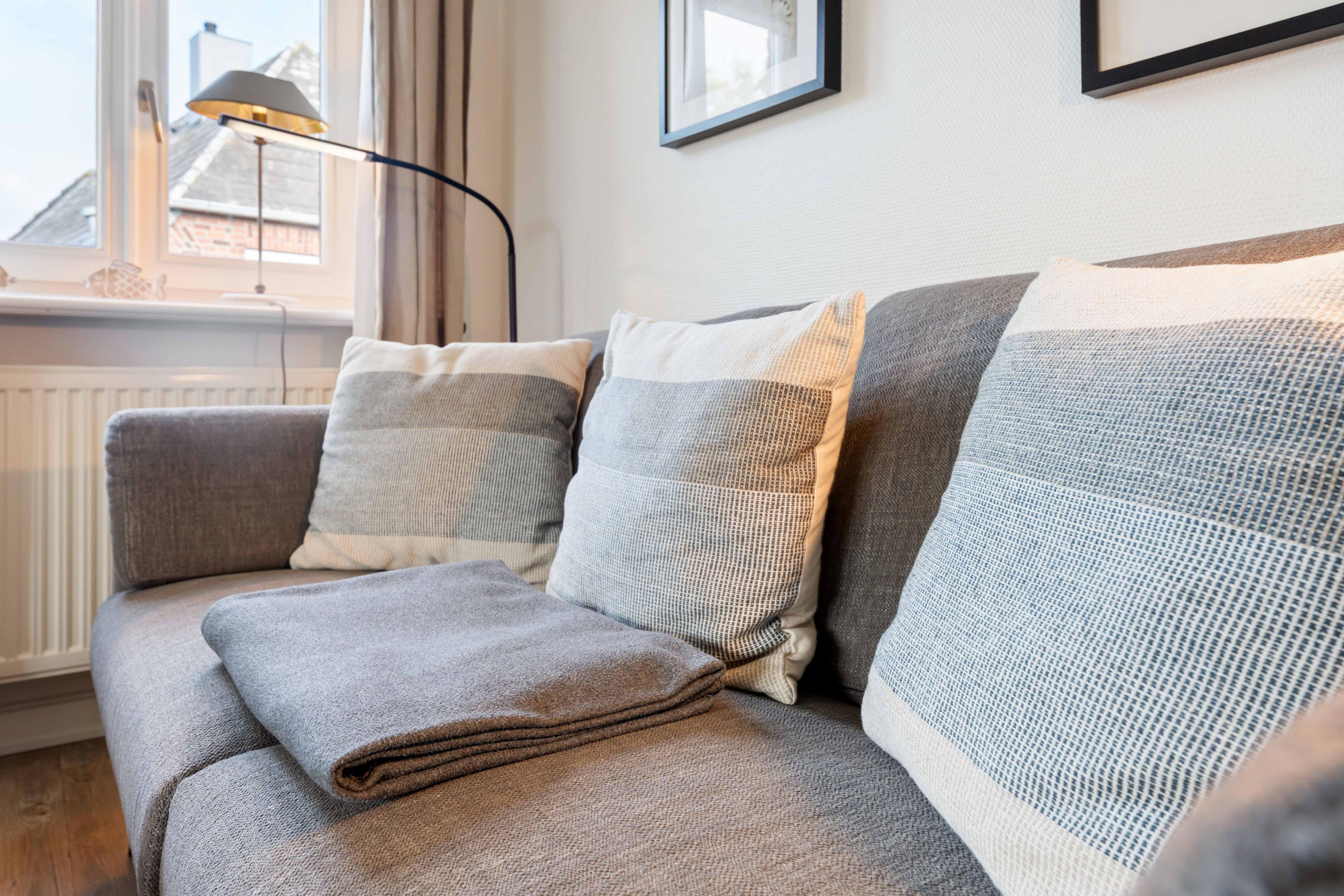 Un canapé gris avec une couverture pliée et deux coussins, un lampadaire et un tableau encadré accroché au mur.