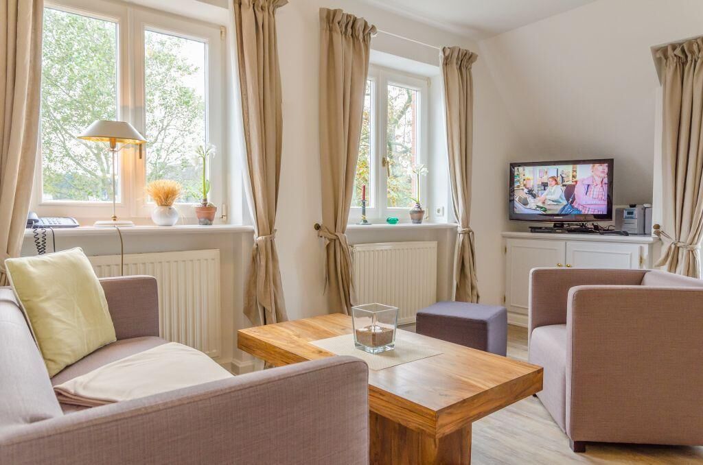 A living room with a sofa, armchairs, a coffee table, a TV, and windows with curtains.