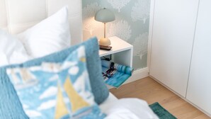 Cama bem arrumada com almofada azul e branca estampada, mesa de cabeceira pequena com abajur, e parede com padrão de árvore.