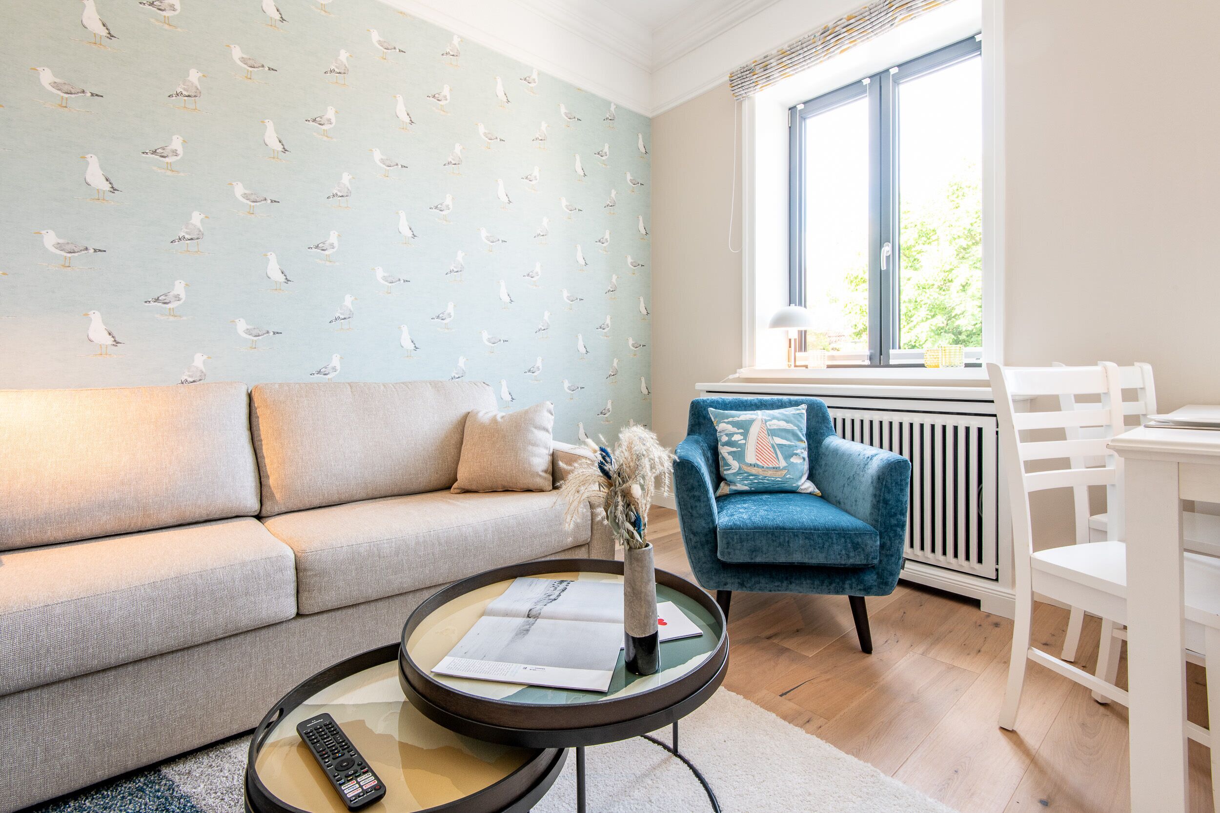 Een woonkamer met een bank, een blauwe fauteuil, een ronde salontafel met een afstandsbediening en een vaas, en een raam met uitzicht op groen.