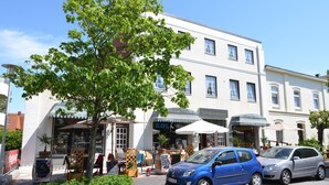 A building with multiple storefronts, including a restaurant and a shop with a sign that reads "SOL". There are two cars parked in front of the building. - Stadthaus Boysenstraße, Whg. 7 Ruhig. Gemütlich. Strandnah. Objekt-id: Shb/07 (Westerland)