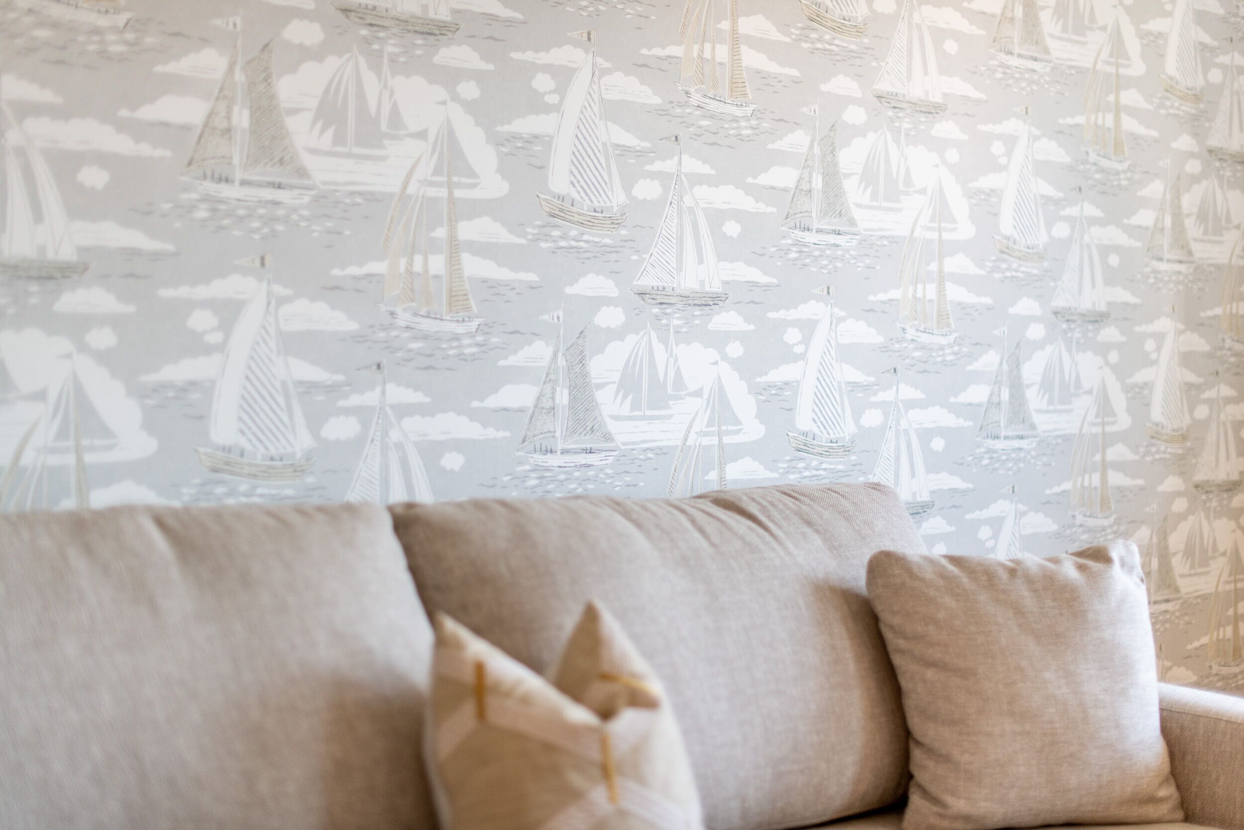 Ein Zimmer mit gestreiftem Wandpapier mit Segelbooten und Wolken, einem beigefarbenen Sofa mit Kissen und einer hellen Wand.