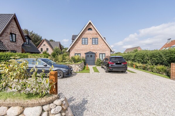 A house with a triangular roof, a gravel driveway, and two parked cars. - Neubau. Balkon. Modern. Süderstraße 42, Whg. Süderholt Objekt-id: Sdw/42 (Westerland)