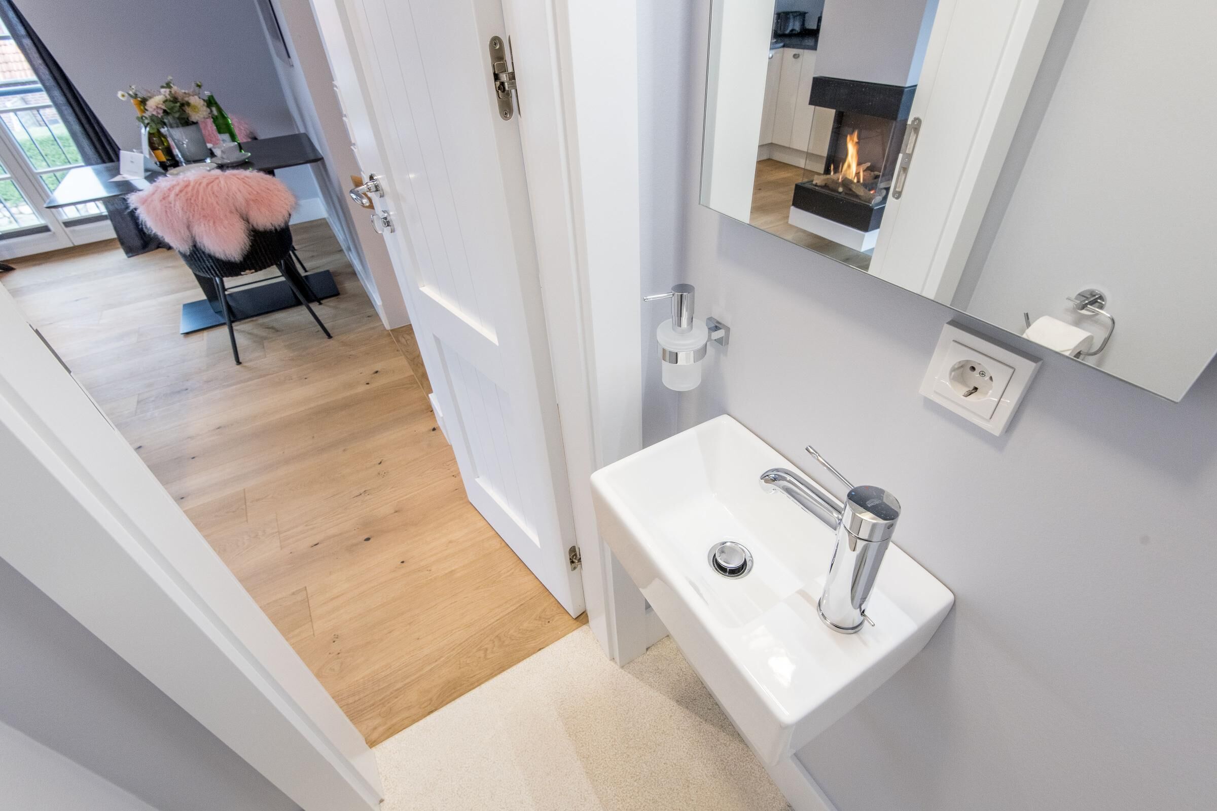 Un baño moderno con un lavabo blanco, un espejo y una puerta que da a un comedor.