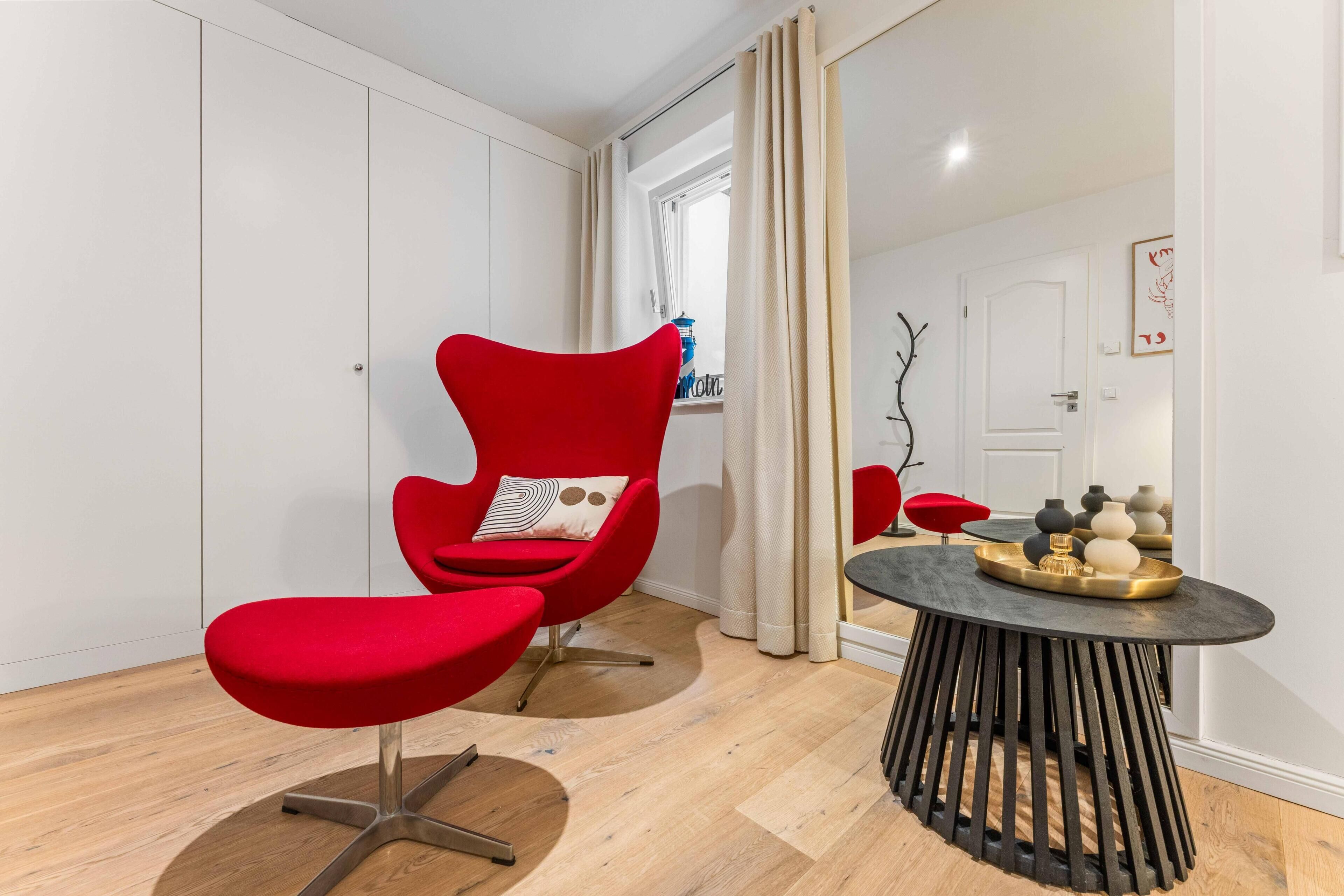 Ein modernes Wohnzimmer mit einem roten Sessel, einem kleinen runden Tisch mit Dekoration und Blick durch ein Fenster mit Vorhängen.