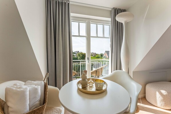 A modern room with a round table, white chairs, a basket with towels, and a view of a balcony with greenery.