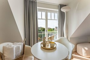 Une chambre moderne avec une table ronde, des chaises blanches, un panier contenant des serviettes et une vue sur un balcon agrémenté de plantes.