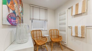 A bathroom with two wicker chairs, a white table, a large painting, and a towel rack. - Stylish. Lichtdurchflutet. Sauna (Westerland)