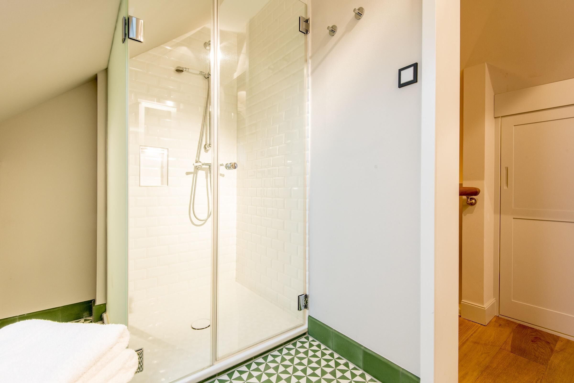 A glass-enclosed shower with a white tiled wall, a green and white patterned floor, and a wooden door in the adjacent room.