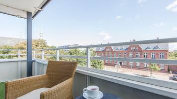 Ein Balkon mit Korbmöbeln und einem Tisch mit Tasse, mit Blick auf ein rotes Backsteinhaus und einen klaren Himmel.