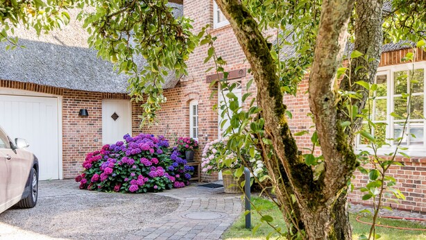 Une maison en briques avec une porte de garage blanche, une allée gravillonnée et un jardin agrémenté de fleurs violettes et roses.