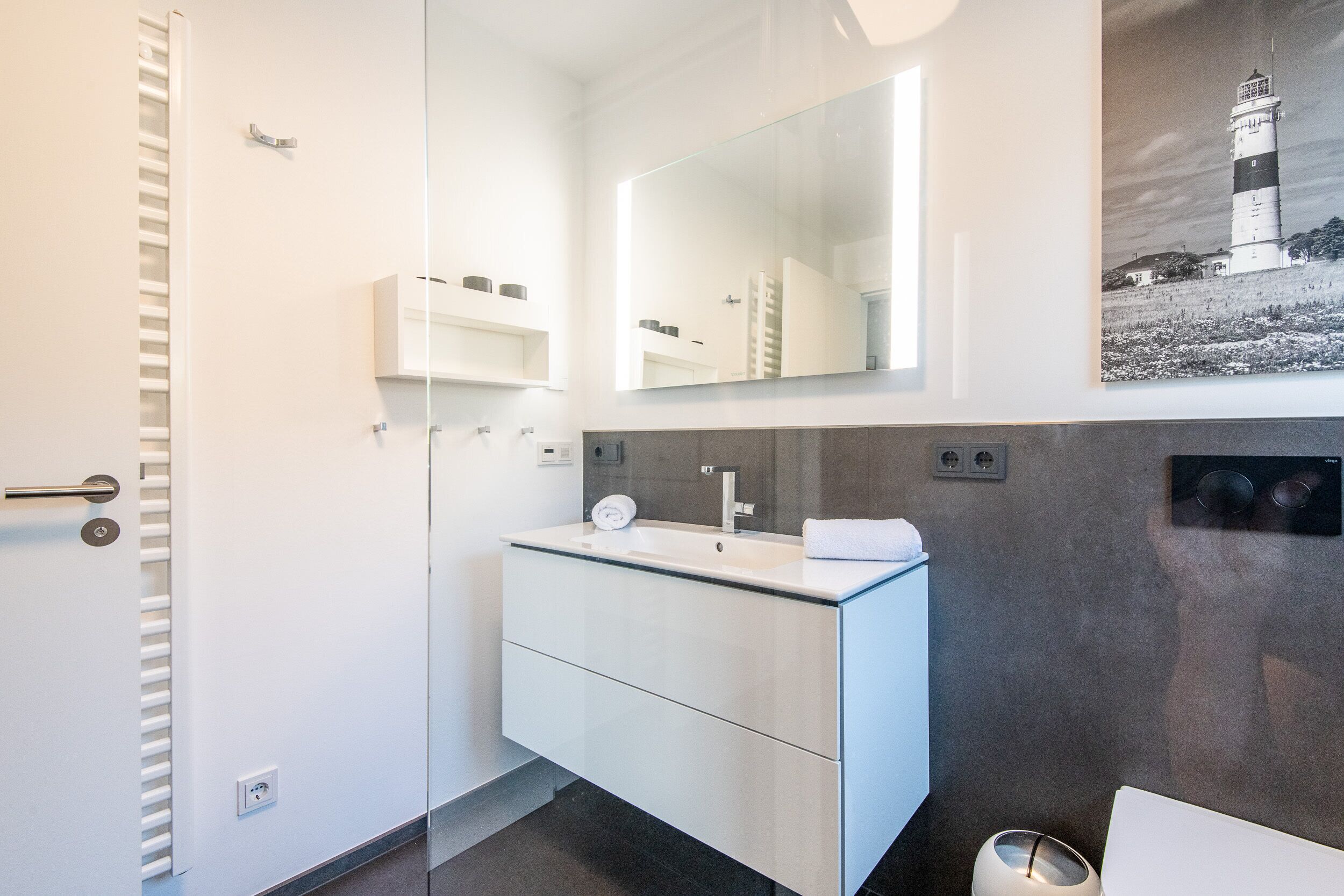 Une salle de bain moderne avec une douche à parois de verre, un meuble-lavabo blanc avec un miroir et une photographie en noir et blanc représentant un phare, accrochée au mur.