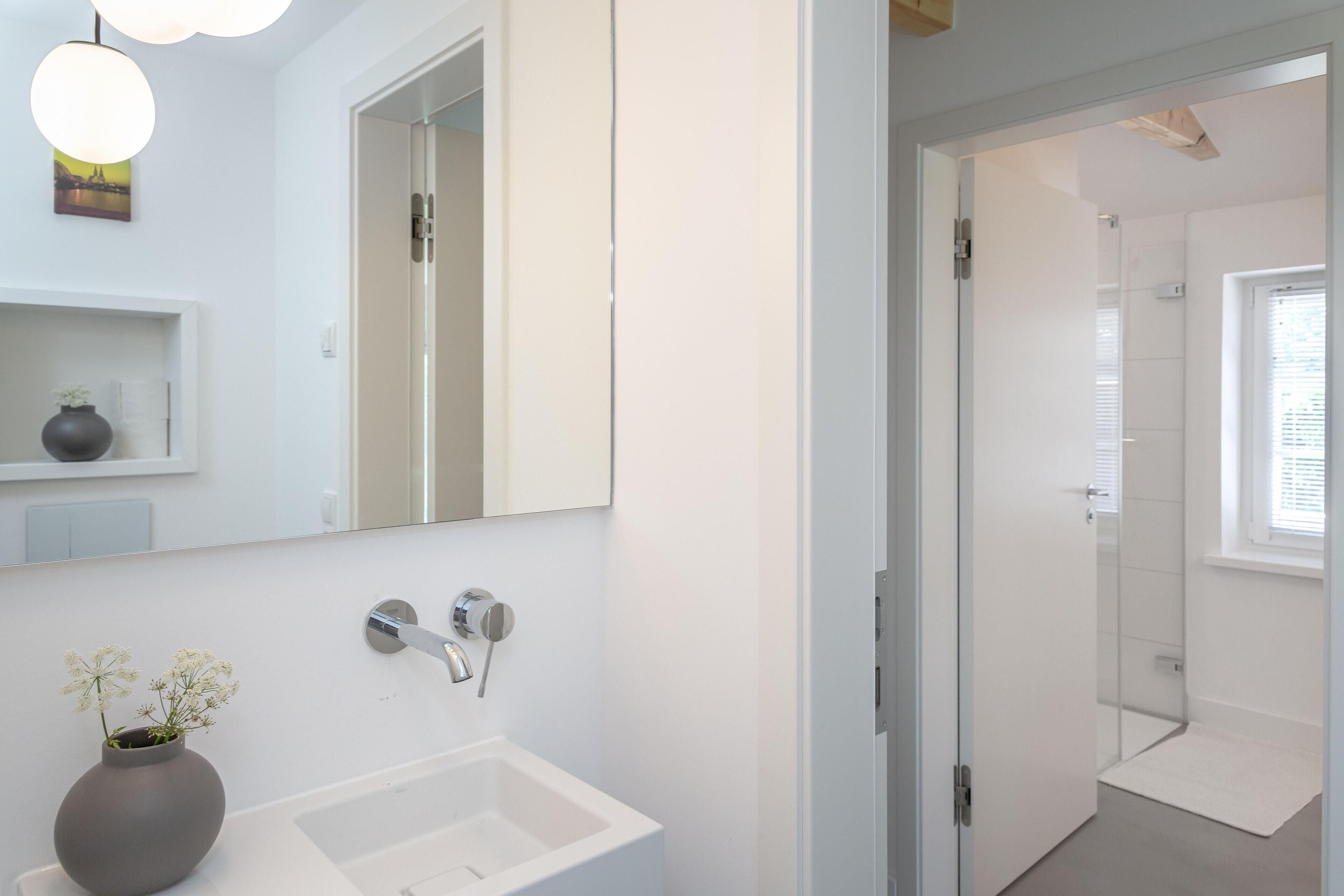 Een moderne badkamer met een witte wastafel, een grote spiegel en een vaas met bloemen.