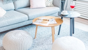 Un soggiorno con un divano grigio, un tavolino in legno con una rivista e due pouf bianchi su un tappeto blu.
