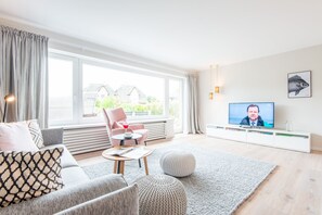 Uma sala de estar moderna com sofá, poltrona e banqueta, uma televisão e vista para o exterior através de grandes janelas.