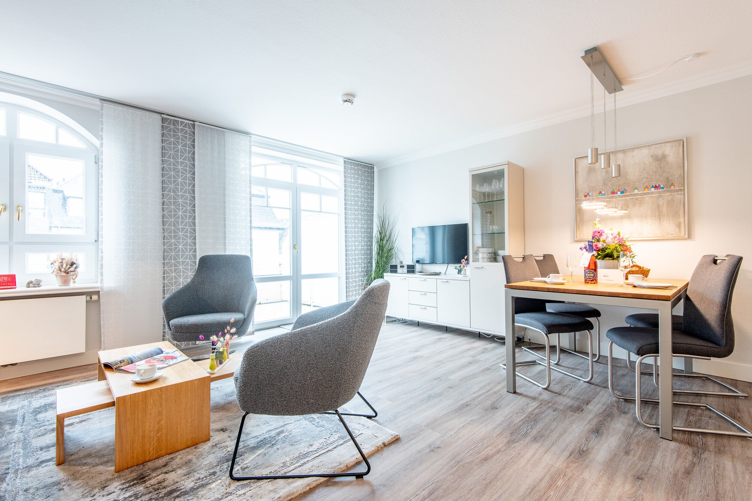 A modern living room with a wooden coffee table, a grey armchair, and a dining area with a wooden table and metal chairs.