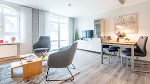 A modern living room with a wooden coffee table, a grey armchair, and a dining area with a wooden table and metal chairs. - Dachterrasse. Stimmig. Zentral. Residenz Bismarck, Whg. 17 Objekt-id: Hrb/17 (Westerland)