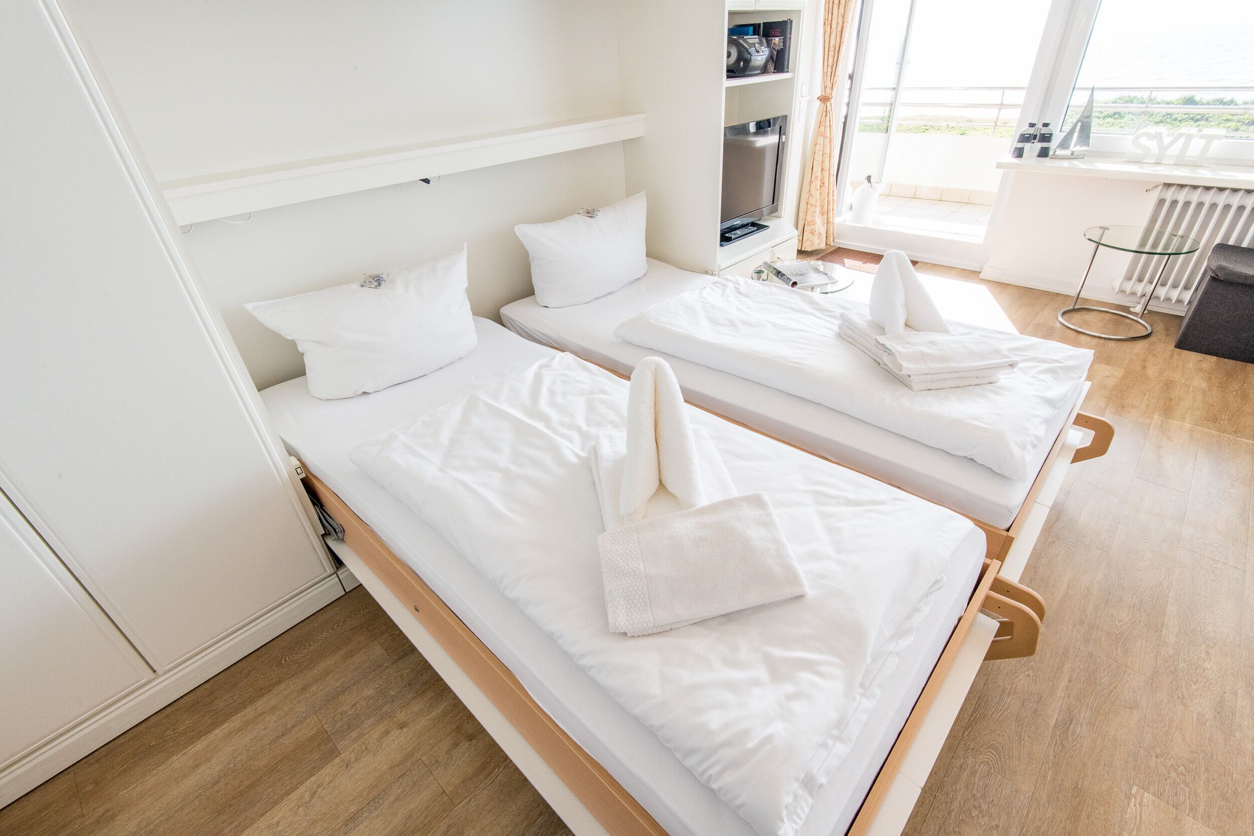Deux lits simples avec du linge de lit blanc, une petite table avec une lampe et une vue sur l’extérieur par une fenêtre.