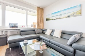 A modern living room with a sectional sofa, a glass coffee table, and a large window with a view of the beach.