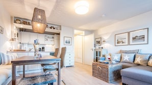 A modern living room with a sofa, a dining table with chairs, and a kitchen area with white cabinets and a hanging light fixture. - Exklusiv. Zentral. Strandnah (Westerland)