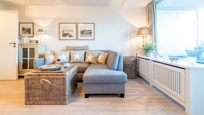 A modern living room with a grey sectional sofa, wicker ottoman, and a large window with a view. - Exklusiv. Zentral. Strandnah (Westerland)