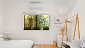A bedroom with a bed, a window with a view of trees, a bedside table, a lamp, a dresser, a chair, and a hanging wooden hanger.