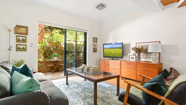 A living room with a sofa, a coffee table, a TV, and a cabinet. There is a sliding glass door leading to a garden.