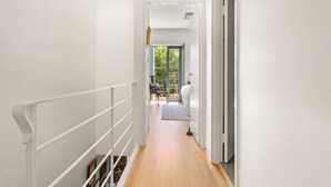 A narrow hallway with white walls and a wooden floor leading to a room with a bed and a chair.