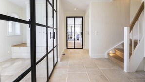 Un pasillo amplio con suelo de baldosas, una puerta de cristal que da a una habitación y una escalera con escalones de madera.