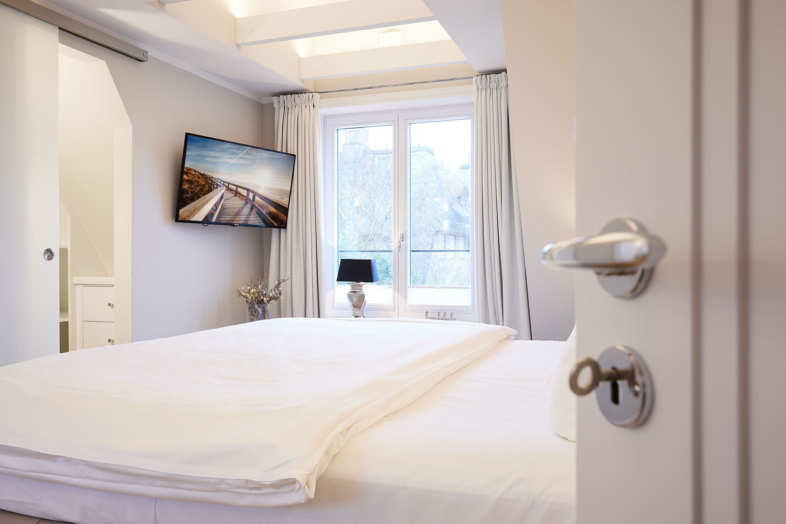 Ein Schlafzimmer mit einem großen Bett, einem Fernseher an der Wand, einem Fenster mit Vorhängen und einer Tür mit Schlüsselloch.