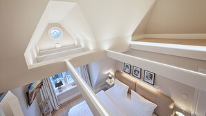 Une chambre avec un plafond en pente, un lit avec une literie blanche, une table de chevet avec une lampe et une fenêtre avec des rideaux.