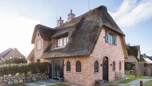 Une maison en briques avec un toit de chaume, un mur en pierre et un petit jardin agrémenté d’un banc.