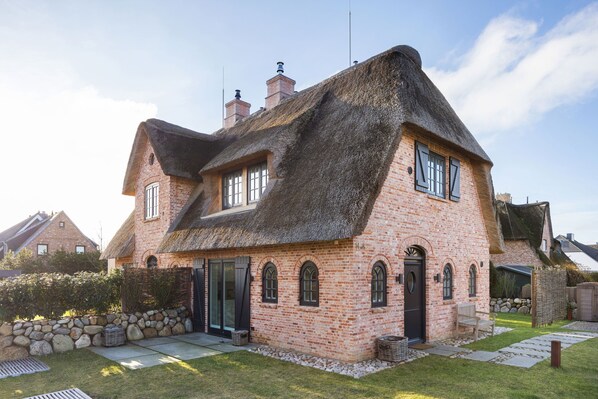 Une maison en briques avec un toit de chaume, un mur en pierre et un petit jardin agrémenté d’un banc.