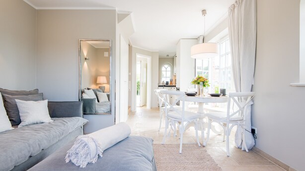 Una habitación de hotel moderna con un sofá gris, una mesa de comedor blanca con sillas y un ventanal con cortinas.
