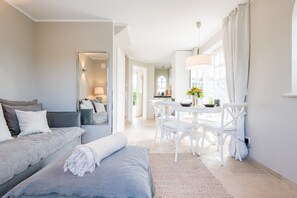 A modern hotel room with a grey sofa, a white dining table with chairs, and a large window with curtains.