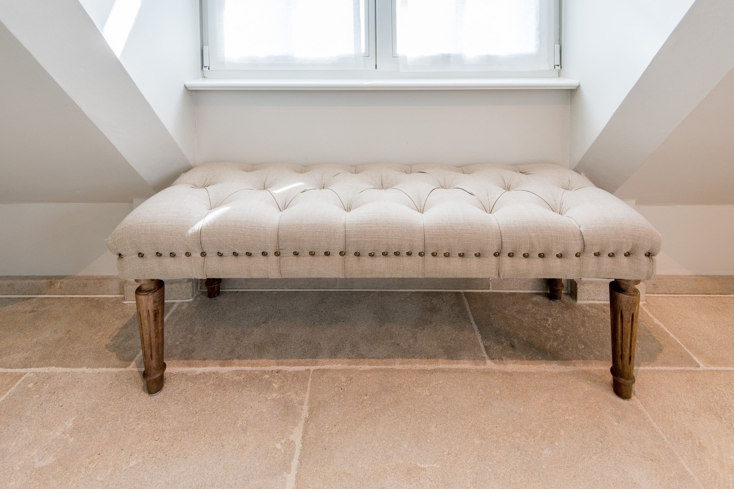 A tufted bench with wooden legs and a cushion, placed under a window with a white frame.