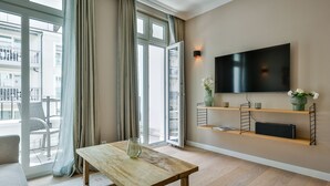 A modern living room with a wooden table, a sofa, a TV mounted on the wall, and a balcony with a view of the city. - Neubau. Strandnah. Anspruchsvoll. Objekt-id: Ljw/05 (Westerland)