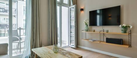 A modern living room with a wooden table, a sofa, a TV mounted on the wall, and a balcony with a view of the city.