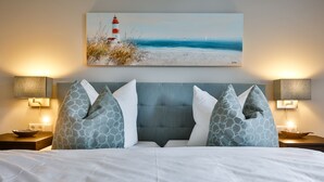 A hotel room with a bed, two nightstands, a painting of a beach scene, and patterned pillows. - Neubau. Strandnah. Anspruchsvoll. Objekt-id: Ljw/07 (Westerland)