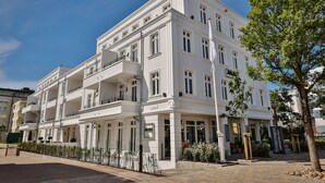 A multi-story building with balconies, a sign indicating 'flintmann', and a paved area with a tree and shrubs. - Neubau. Strandnah. Anspruchsvoll. Objekt-id: Ljw/11 (Westerland)