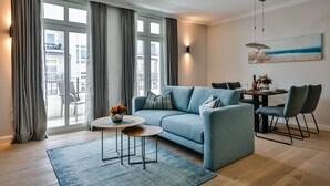 Una sala de estar moderna con un sofá, una mesa de comedor y sillas. Hay un ventanal con cortinas, un cuadro en la pared y una mesita con flores.
